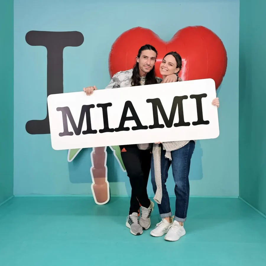 Celebrando-el-amor-en-Miami Pareja sonriente sosteniendo un cartel de "I ❤️ Miami" en un fondo azul, transmitiendo alegría y amor por la ciudad.